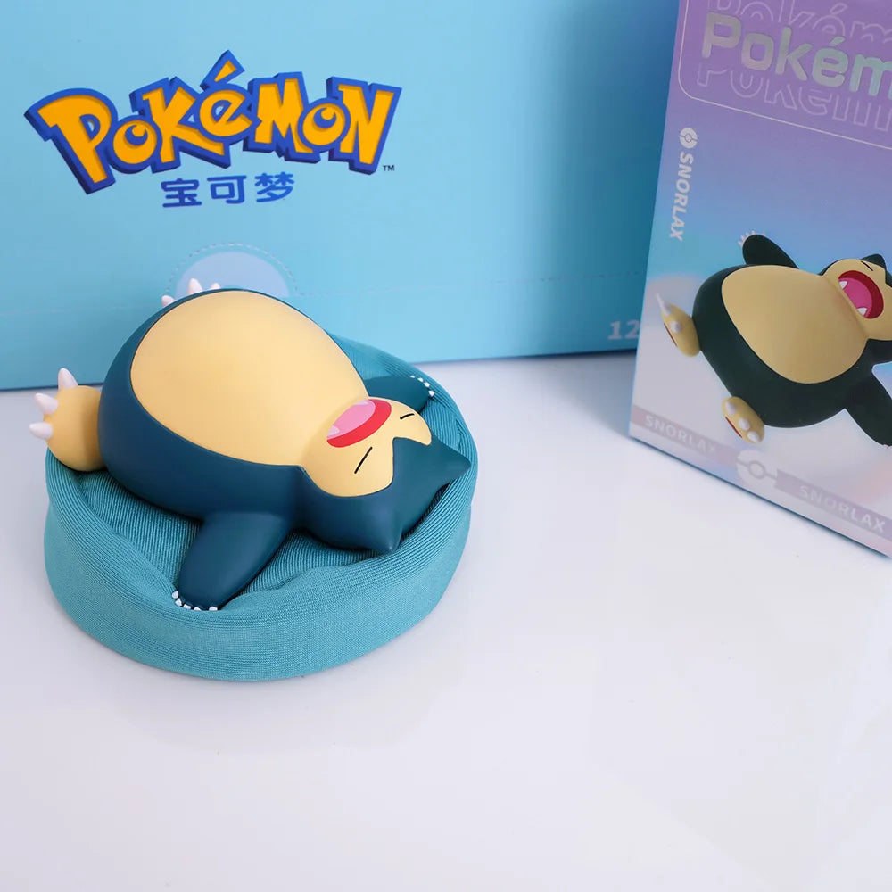 Snorlax figurine sleeping on a teal/blue cushion/pillow, featuring a relaxed design, with a Pokémon-themed box and branded background.