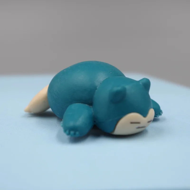 Tiny Snorlax figurine lying in a sleeping position on a light blue surface, showcasing its adorable and compact design.