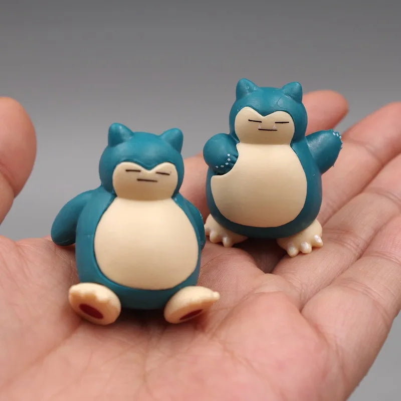 Two tiny Snorlax figurines displayed on a hand, one sitting and one waving, highlighting their small size and detailed design.